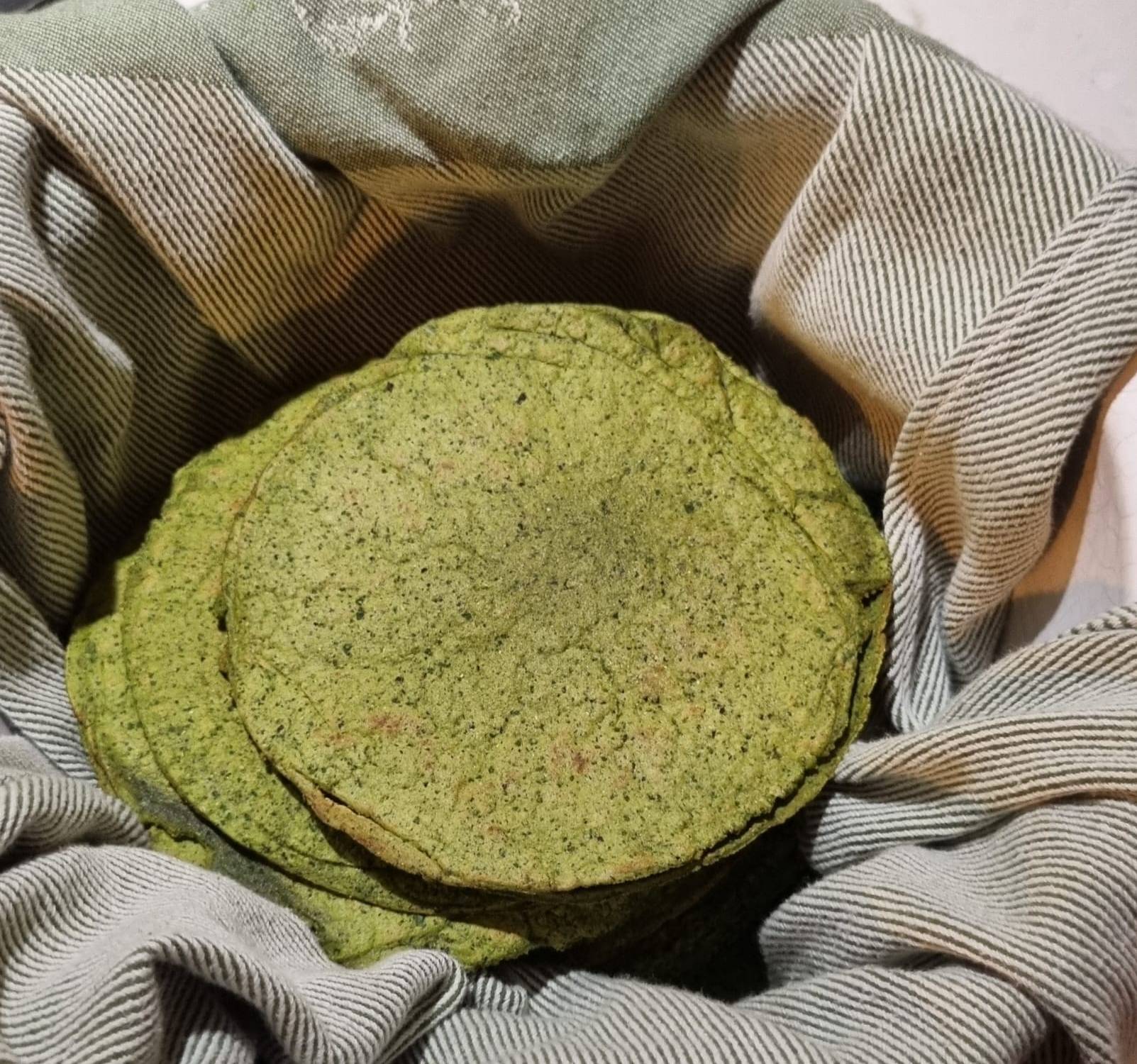 A stack of homemade spinach and beans tortillas with golden-brown spots, slightly charred edges, and a soft texture. The tortillas have a subtle green hue from the spinach and are neatly arranged on a plate with a cloth partially covering them to keep them warm.