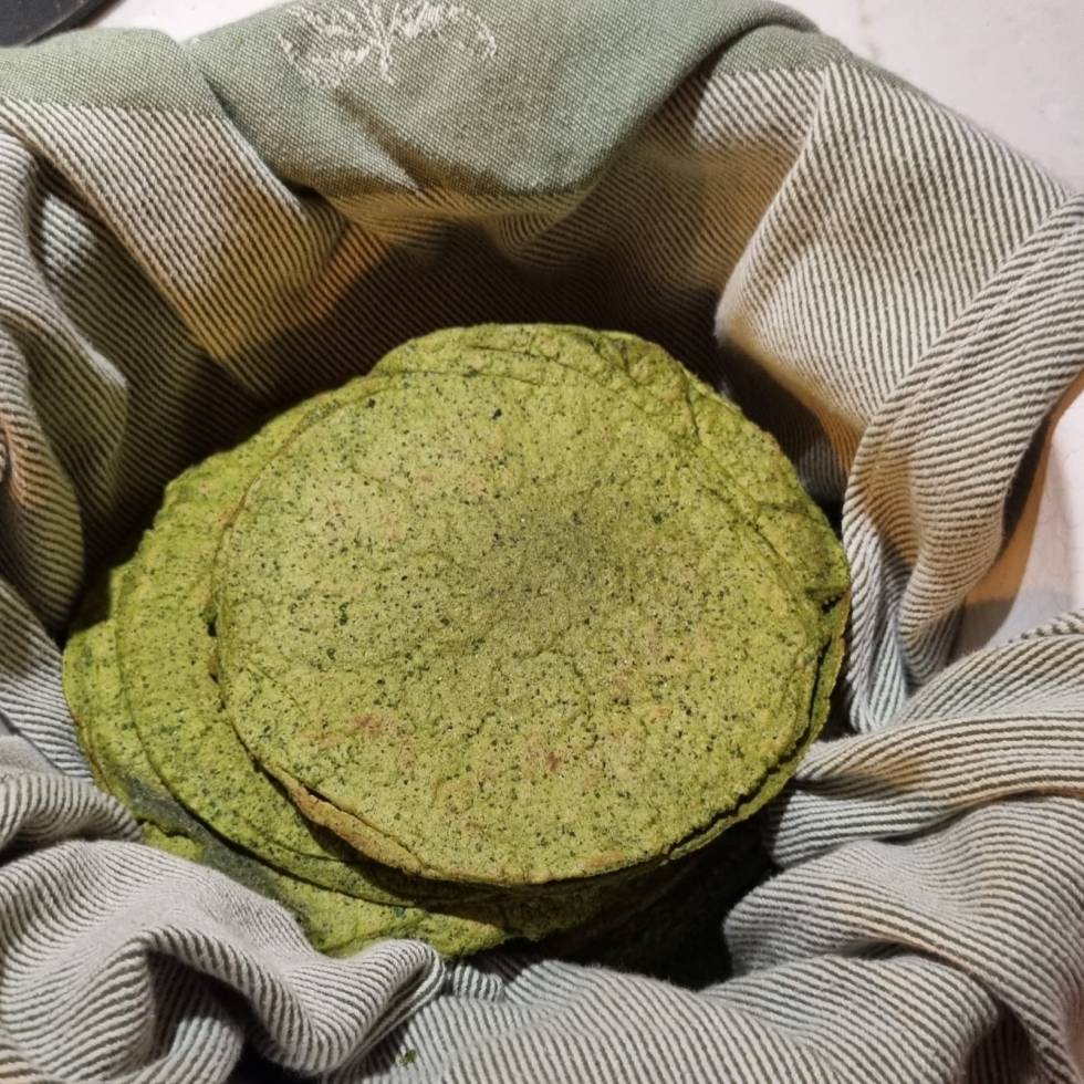 A stack of homemade spinach and beans tortillas with golden-brown spots, slightly charred edges, and a soft texture. The tortillas have a subtle green hue from the spinach and are neatly arranged on a plate with a cloth partially covering them to keep them warm.