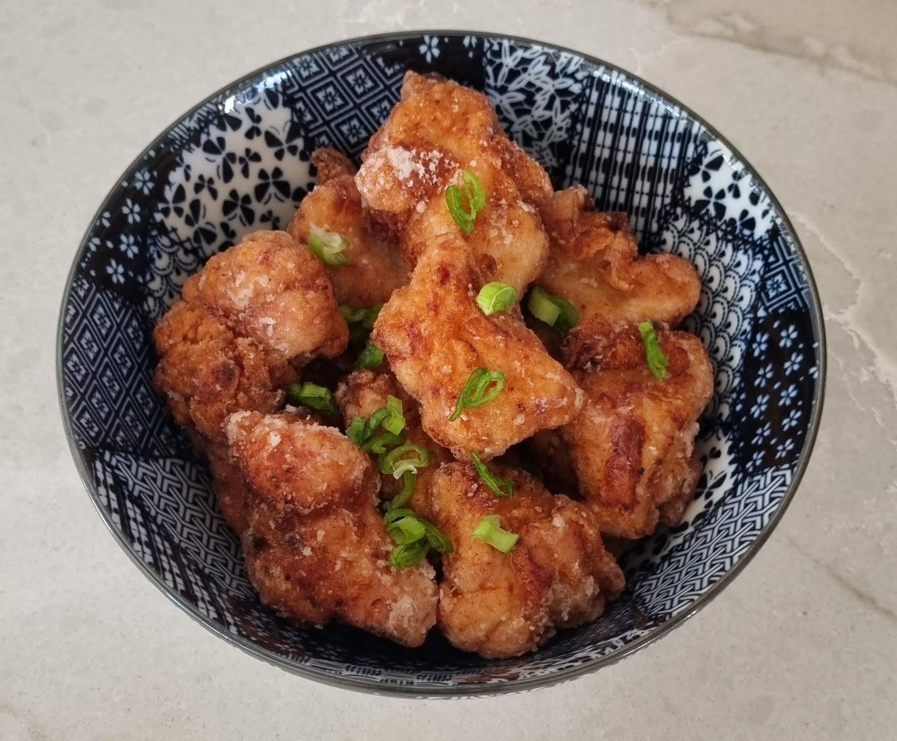 A bowl of golden-brown Japanese fried chicken (karaage) garnished with sliced green onions, served in a black and white patterned ceramic bowl on a light marble countertop.