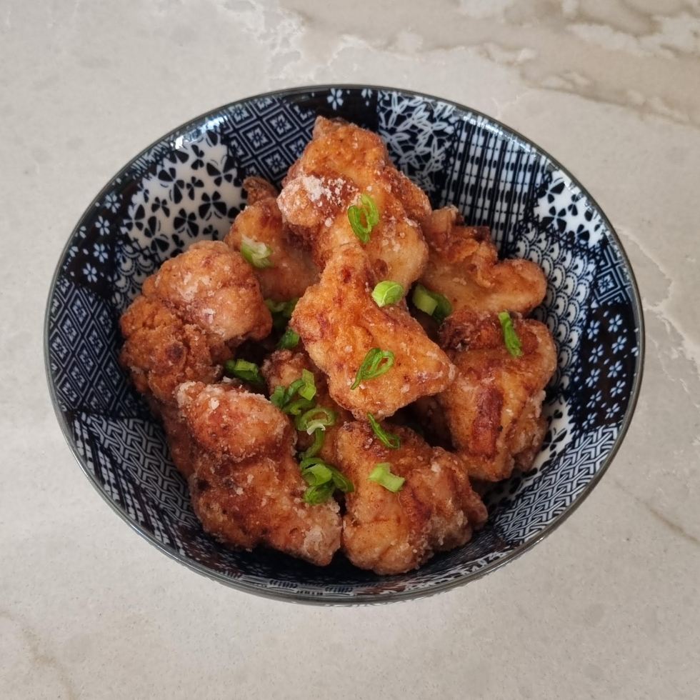 A bowl of golden-brown Japanese fried chicken (karaage) garnished with sliced green onions, served in a black and white patterned ceramic bowl on a light marble countertop.