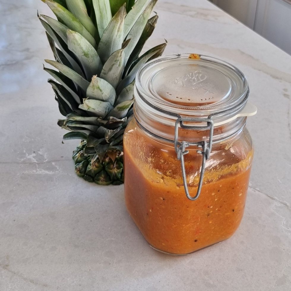 Glass jar filled with vibrant orange-red pineapple hot sauce sitting next to a leafy green pineapple top on a white marble countertop.
