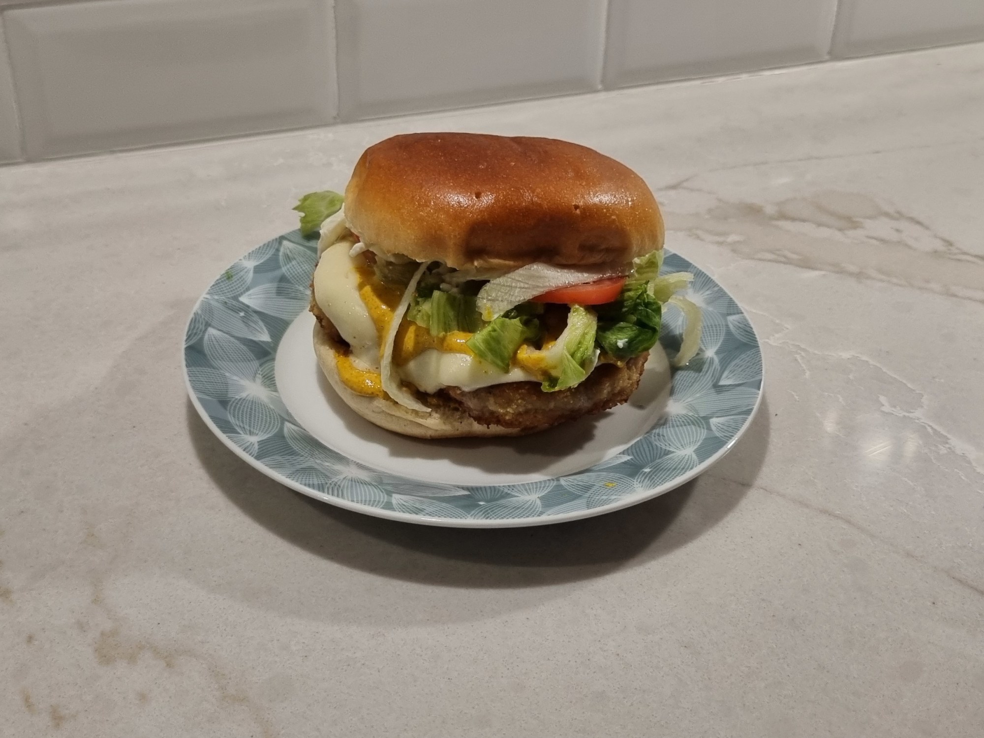 A homemade chicken curry burger on a small patterned plate, topped with melted cheese, yellow curry sauce, lettuce, sliced tomato, and onions inside a toasted bun.