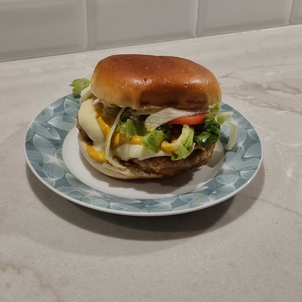 A homemade chicken curry burger on a small patterned plate, topped with melted cheese, yellow curry sauce, lettuce, sliced tomato, and onions inside a toasted bun.